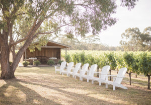 A beautiful outdoor wedding ceremony setup among green vineyards in Swan Valley, with white chairs and floral decorations under a glowing sunset.