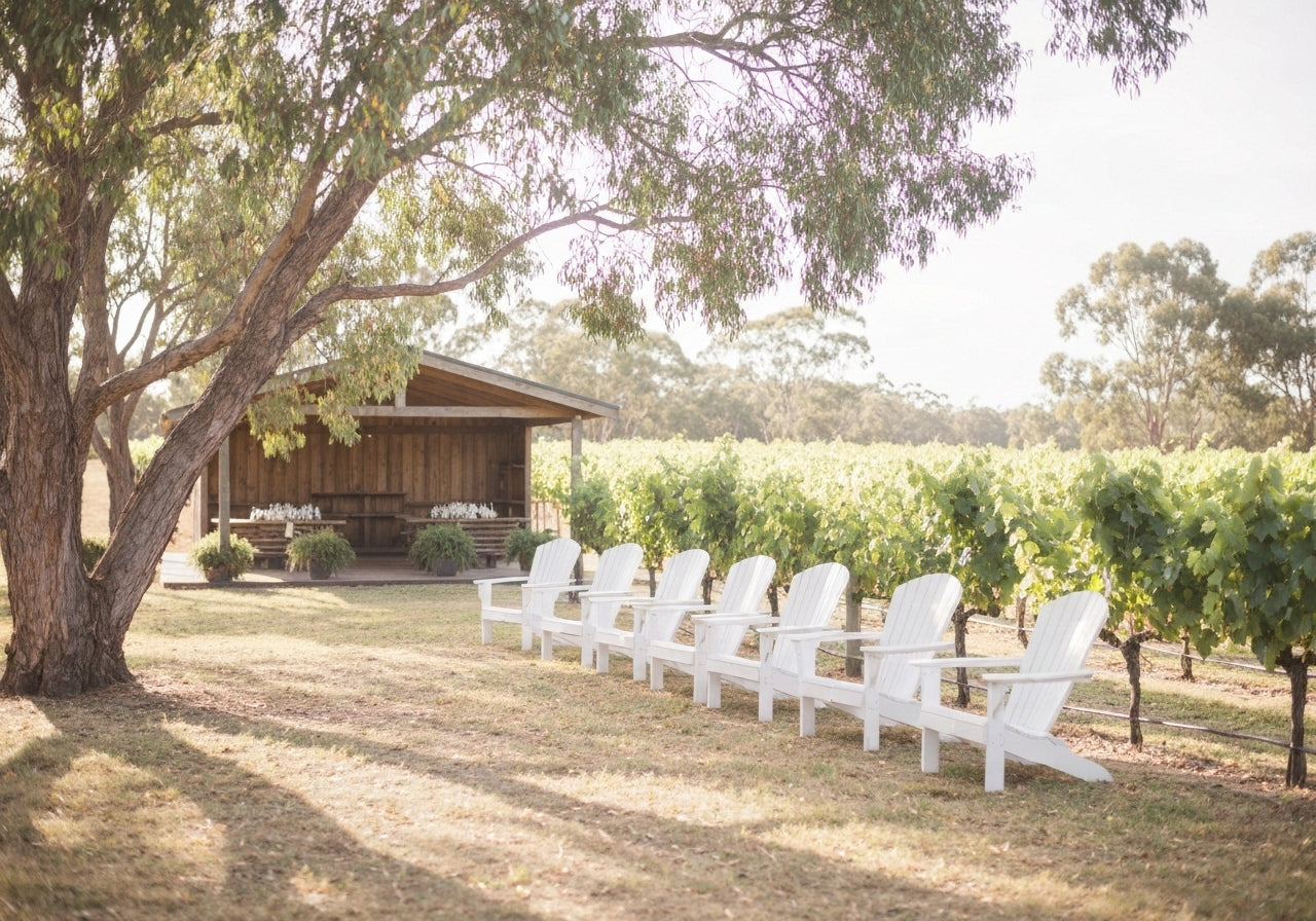A beautiful outdoor wedding ceremony setup among green vineyards in Swan Valley, with white chairs and floral decorations under a glowing sunset.