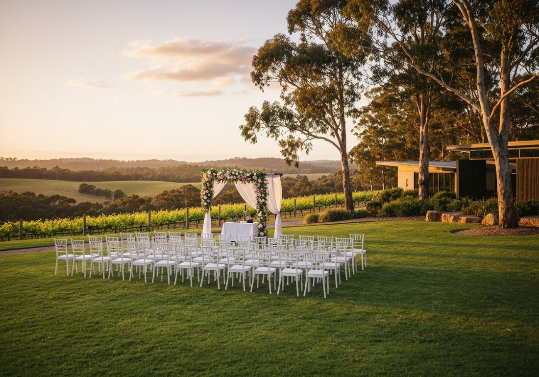 A picturesque outdoor wedding setup in the Margaret River Region, featuring a floral arch, white chairs, vineyards, and a winery building at sunset.