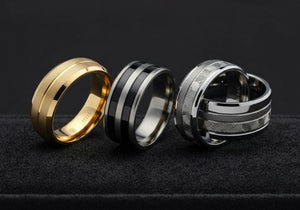 Various men's wedding rings made from gold, platinum, titanium, tungsten, and ceramic displayed on a reflective surface, showing different styles and material characteristics.