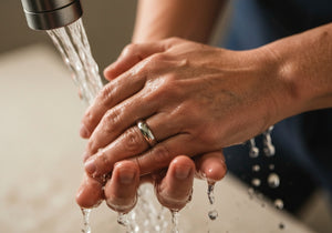 Close-up of hands with a wedding ring being washed carefully under running water in a bathroom sink.