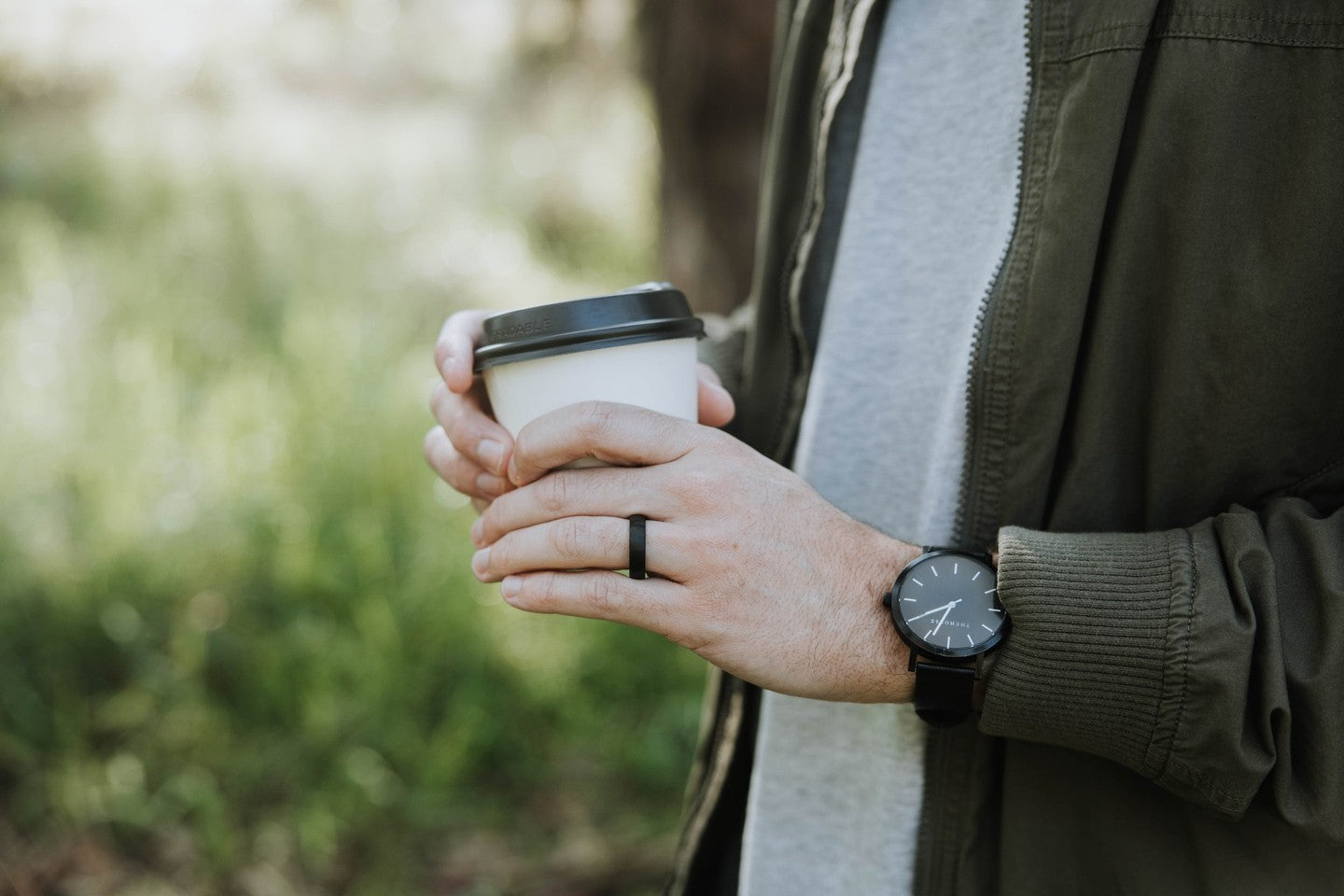 Person wearing a black tungsten wedding ring while holding a coffee cup outdoors with a blurred natural background