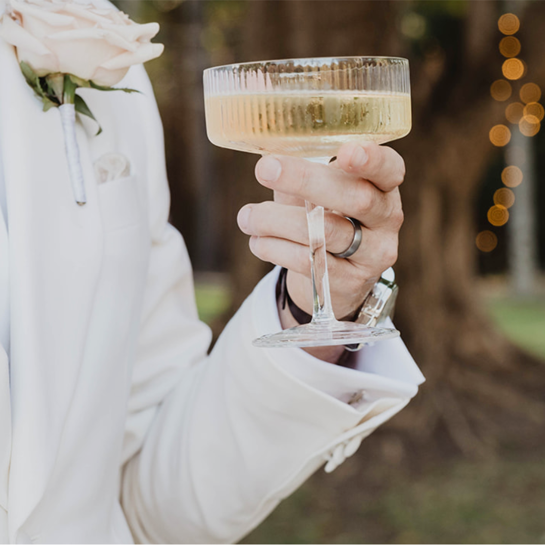 Person holding a cocktail glass with a blurred background while wearing a tungsten wedding ring.