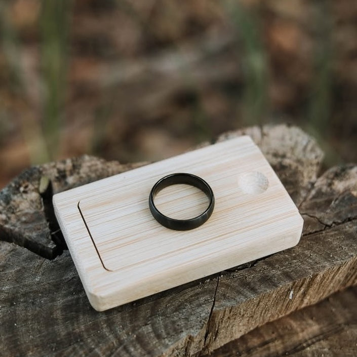 Wooden ring box with a black wedding ring on top.