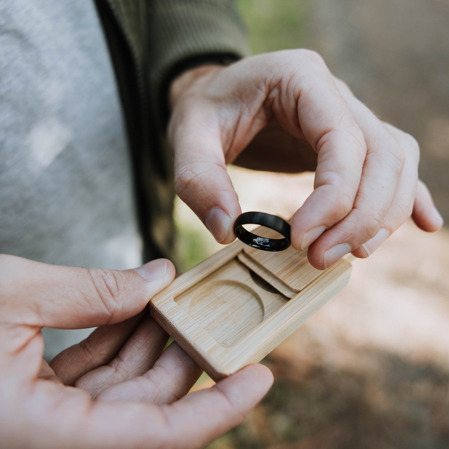 Two hands holding a wooden ring box with a black wedding band inside, outdoors.